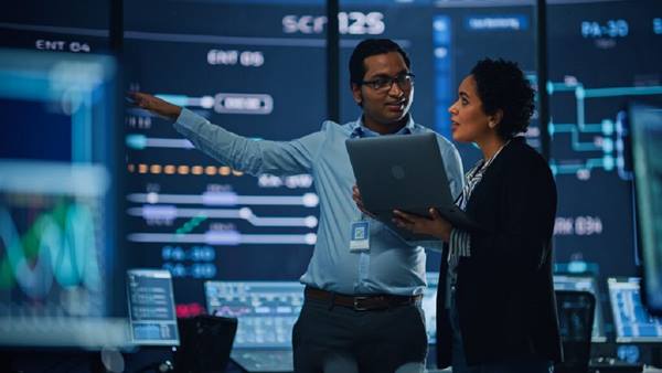 Conversation among professionals with multiple monitors displaying graphics, highlighting solutions to assess and predict population risk with advanced analytics and SaaS tools.